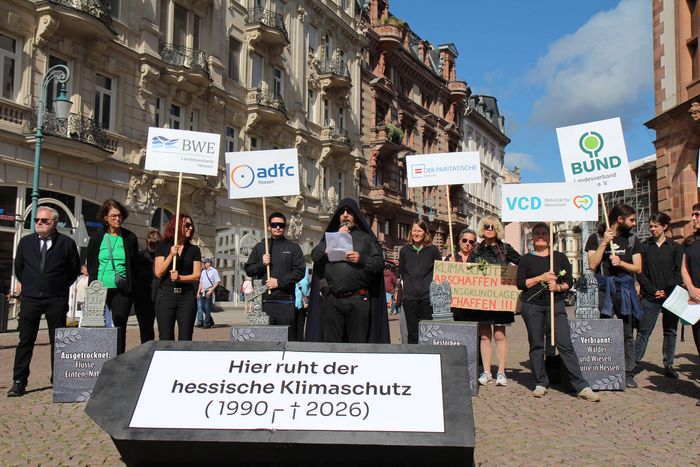 Ausschnitt aus der Protestaktion: Die Gäste der Beerdigung stehen hinter dem Trauerredner (Dr. Werner Neumann) und halten Schilder der Organisationen hoch, die hinter der Protestaktion stehen.