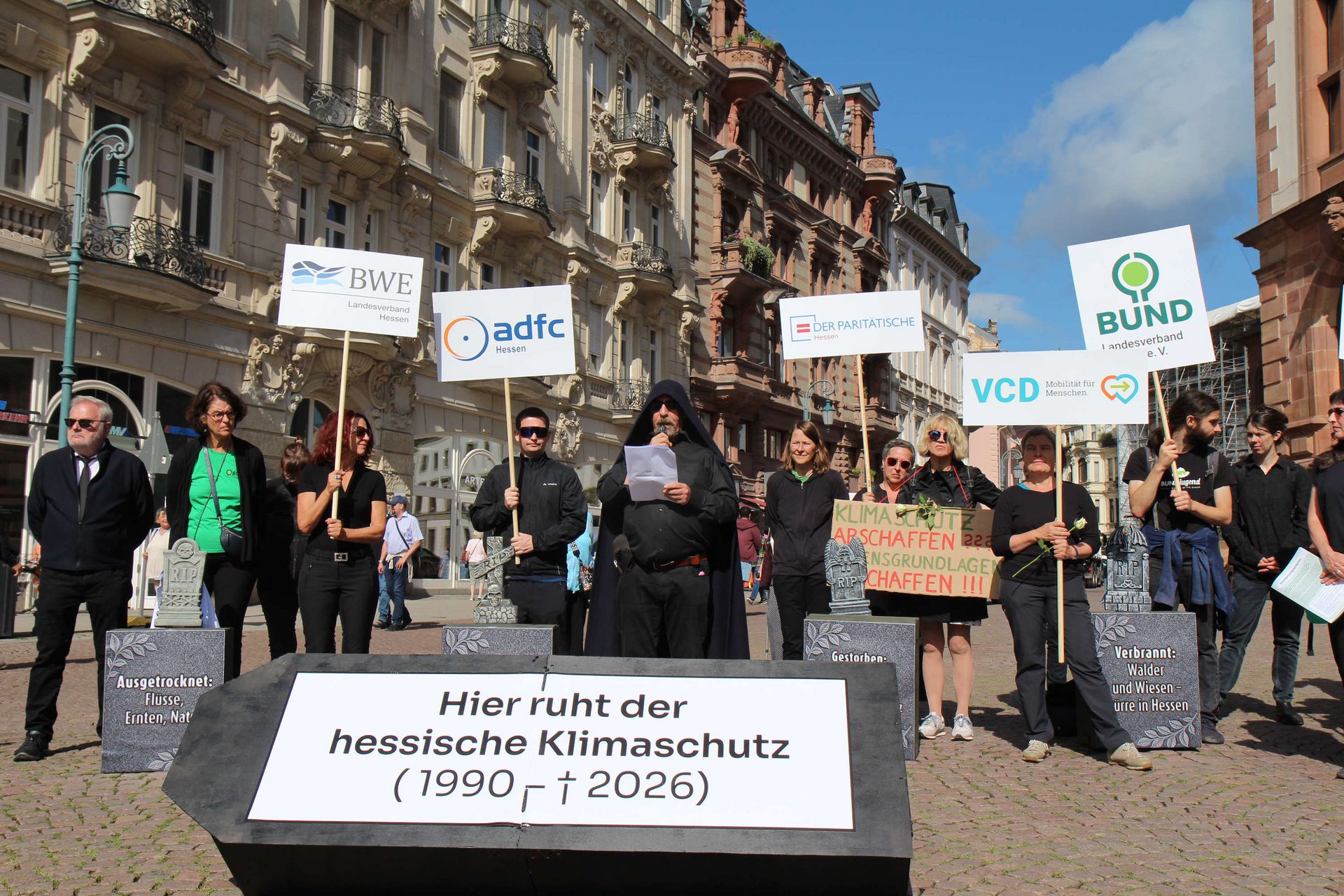 Ausschnitt aus der Protestaktion: Die Gäste der Beerdigung stehen hinter dem Trauerredner (Dr. Werner Neumann) und halten Schilder der Organisationen hoch, die hinter der Protestaktion stehen.