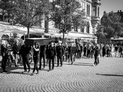 Vier Menschen tragen einen Sarg. Ihnen folgen wiederum weitere Menschen, eine Trauergemeinde. Das Bild ist in Schwarz-Weiß.
